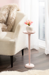 Tall, slender pale pink side table with a small vase of pink flowers, positioned next to a white upholstered armchair with a faux fur throw in a bright room.