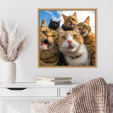 Framed wall art showing a close-up selfie of five curious cats with varied tabby and black fur against a blue sky background, displayed above a white desk.