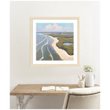 Coastal landscape painting showing a sandy shoreline curving alongside blue waves and marshy green wetlands under a pastel sky, framed on a white wall above a desk.