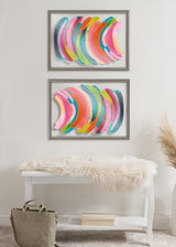 Two framed abstract prints by Jessi Raulet of Ettavee featuring curved brushstrokes in bright pink, green, yellow, and blue on a white wall above a white bench with a fur throw and a gray bag.