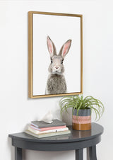 Framed canvas print of a young rabbit with upright ears against a white background, hanging above a gray half-moon table with stacked books, a bird figurine, and a potted plant.