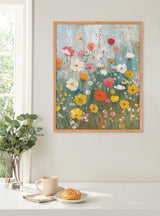 Painting of a wildflower garden with red, yellow, orange, and white flowers on a light blue background, framed in light wood, hanging on a white wall above a table.