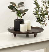 Dark brown decorative tray with a wavy edge and three short legs holding a black planter with green leaves and two white pillar candles on a white surface.
