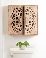 Square wall cabinet with intricately carved floral and circular wood patterns on two doors, mounted above a white toilet tank with a small green potted plant.