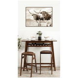 Sepia-toned framed canvas print of two Texas Longhorn cattle in a pasture hanging above a wooden bar table with two matching stools and a small potted plant.
