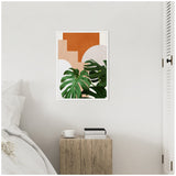Abstract botanical framed canvas with two large green monstera leaves overlaying geometric shapes in terracotta and beige, hanging on a white wall above a textured wooden side table.