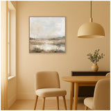 Neutral abstract landscape painting with soft gray, beige, and brown tones in a thin silver frame, hanging above a wooden dining table with tan chairs and a beige pendant light.