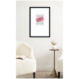 Framed print of a pink ticket with text 'One Life' over grayscale newspaper background, hanging on a white wall between two cream armchairs and a small round table.