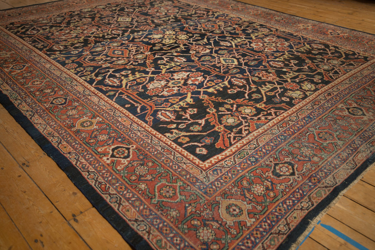 Vintage Mahal carpet with intricate floral and vine patterns in muted reds, greens, and blues on a dark central field, bordered by geometric designs, laid on wooden floorboards.