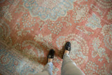 Worn Mahal carpet in muted red and faded blue with intricate floral and geometric patterns, seen from above with feet in black loafers and light pants standing on it.