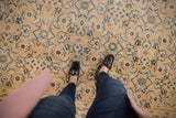 Top-down view of a person wearing dark jeans, black leather shoes, and a pink coat standing on a beige vintage Mahal carpet with blue floral patterns.