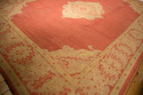 Vintage Oushak carpet with a large faded red central field featuring a beige medallion and a wide beige border decorated with red floral patterns, laid on a wooden floor.