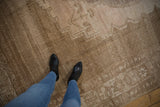 Person wearing blue jeans and black boots standing on a large vintage distressed Oushak carpet in muted beige and brown tones with intricate faded patterns.