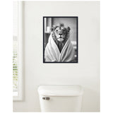 Black and white framed canvas of a lion wrapped in a towel, hanging on a white bathroom wall above a white toilet.