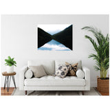 Minimalist framed canvas showing mountain silhouettes reflected symmetrically on a calm lake at sunrise, hung above a white sofa with neutral and patterned pillows in a bright room.