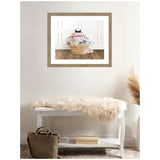 Framed photo of a wicker laundry basket overflowing with clothes and a black cat peeking from inside, hung above a white bench with a cream fur throw.