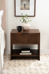 Dark brown wood side table with chevron slats on the drawer front, a black mug and striped ceramic vase on top, and books on the open shelf below.