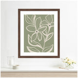 Framed wall art print depicting delicate cream-colored flowers with visible veins on a muted green background in a dark brown frame hung above a white shelf with glassware and small flower arrangement.
