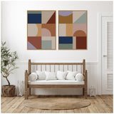 Two framed geometric abstract prints above a wooden bench with white cushions, featuring earthy tones and muted blues in a minimalist room with a round woven rug and a potted plant.