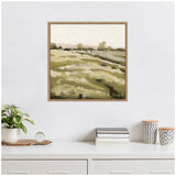 Framed canvas print of a landscape with muted green and beige fields, distant trees, and a pale sky, hung above a white desk with plants and books.