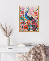 Framed colorful print of a blue peacock surrounded by pink, orange, and red flowers with green leaves on a white wall above a white desk with books and a vase of dried grasses.