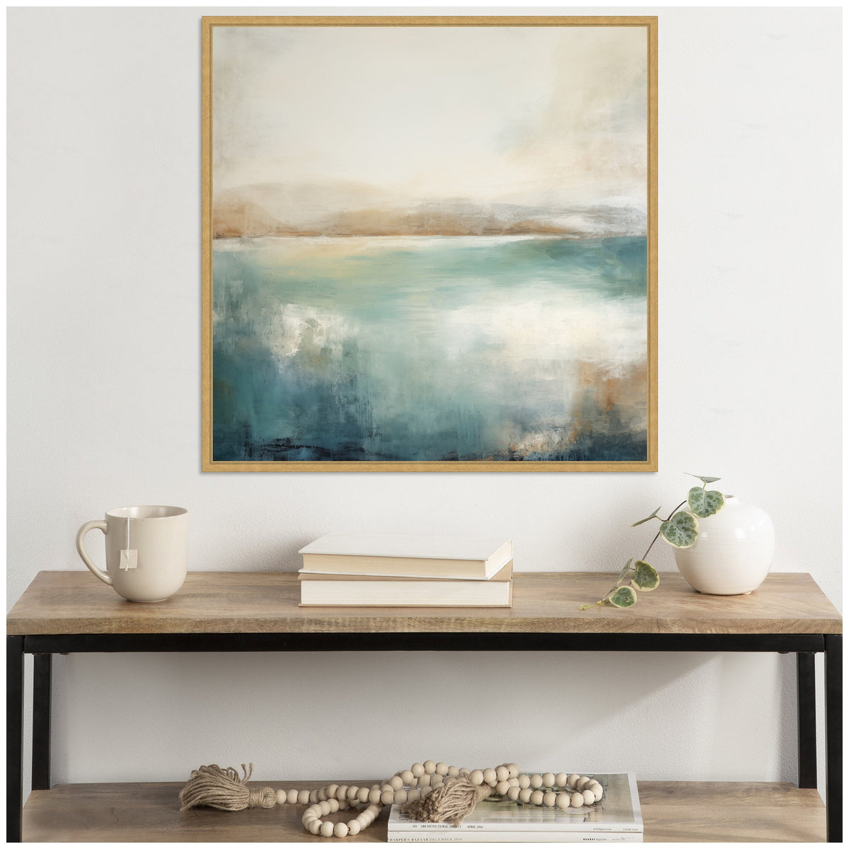 Abstract lake landscape artwork with layered teal, beige, and white hues in a light wood frame above a rustic wood and black metal console table with a beige mug, books, and a white vase with a trailing plant.