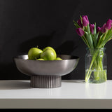 Brushed metal bowl with vertical ribbed base filled with green apples, beside a clear glass jar of purple tulips on a white surface against black background.