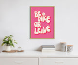 Framed canvas with bold, rounded cream letters outlined in pink reading 'Be Nice or Leave' on a bright pink background, hanging above a white dresser with plants and stacked books.