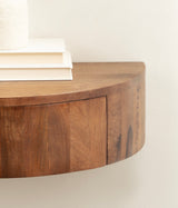 Close-up of a curved, wall-mounted floating wood side table with smooth grain and a warm brown finish against a light wall, holding stacked white books.