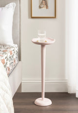 Tall, slender pink Alessia Colored Drink Table with a shallow round top holding a glass of water, positioned beside a bed with floral bedding in a bright bedroom corner.