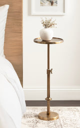 Adjustable polished brass metal side table with a round top holding a white vase with dried flowers, placed beside a bed with white bedding and a wooden headboard.