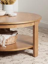 Round wooden coffee table with light natural finish, featuring a lower shelf holding a woven basket with a beige throw, placed on a textured light rug.