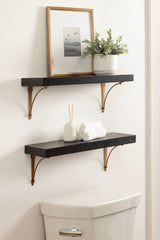 Two black floating shelves with brass brackets mounted on a white wall above a white toilet, holding a framed artwork with birds and a small potted plant on the top shelf and rolled white towels with a diffuser on the bottom.
