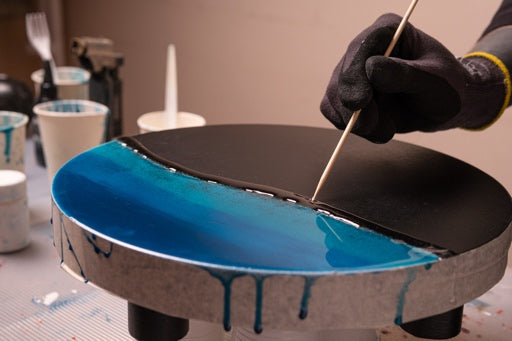 Person applying blue paint to a round object using a stick, with a blurred background of art supplies.