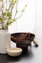 Dark bronze ribbed metal bowl with a wooden bead necklace inside, displayed on a black surface next to a white vase with green branches and a small beige candle dish.