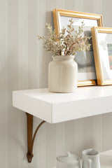 White floating shelf supported by a simple wooden bracket, displaying a cream ceramic vase with dried flowers and two gold-framed pictures leaning against a striped wallpaper.