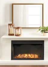 Pair of cylindrical wooden and glass candle holders with white candles on a white mantel above a black-framed electric fireplace with glowing embers, next to a green plant in a pot.