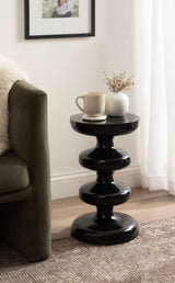 Glossy black metal side table with three rounded stacked tiers, placed next to a green velvet sofa with a white throw in a softly lit room.