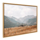 Framed canvas showing a wide valley with golden grass foreground and layered mountain ranges under a cloudy sky.