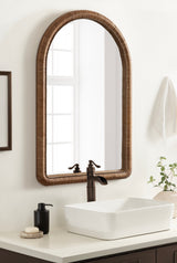 Arched wall mirror with textured brown beaded frame above a white rectangular sink and dark faucet, set against a light wall with minimal decor.