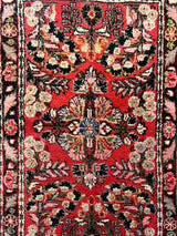 Small Sarouk rug section with red background, floral motifs in beige, black, cream, and blue, framed by intricate black and pink borders.
