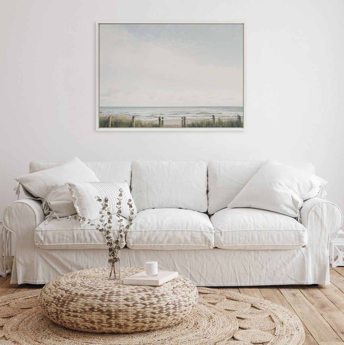 Minimalist living room with a white cushioned sofa, a textured woven round ottoman holding a book and vase, and a pale seaside landscape framed artwork above.