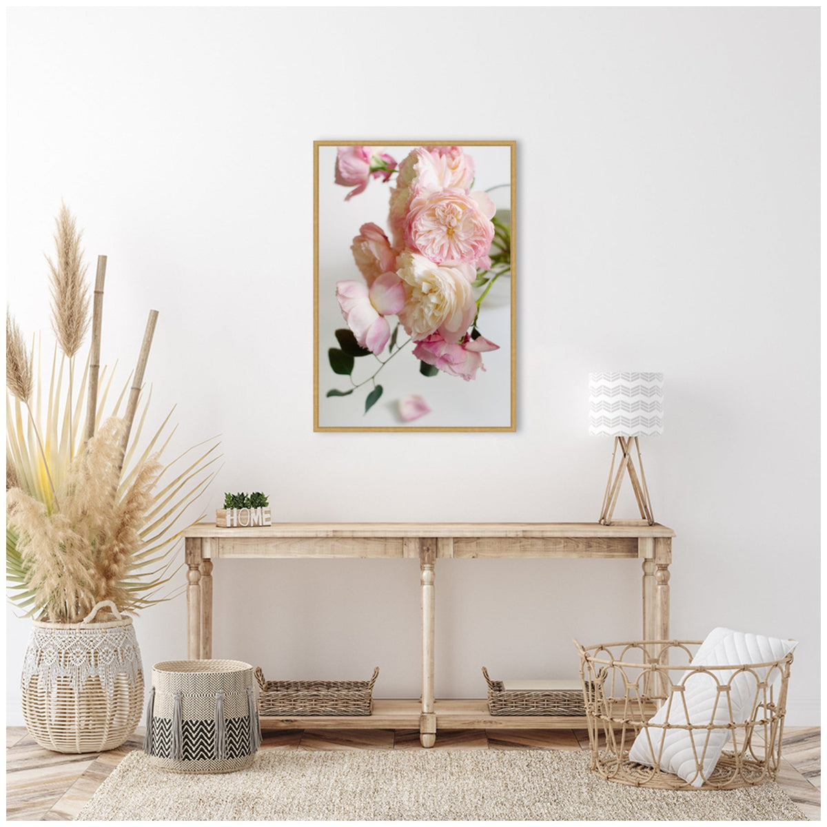 Soft pink and cream roses with green leaves on a white background in a natural wood frame hanging above a rustic wooden console table with neutral woven baskets and pampas grass.