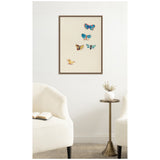 Framed canvas print of five colorful butterflies in blue, yellow, and orange tones on a cream background, hanging on a white wall above a small brass side table.