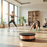 Glossy black and copper circular Qi-Home Cell device on wooden floor with blurred group doing yoga poses in bright studio background