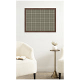 Sage green fabric pinboard with white plaid grid pattern framed in dark wood, mounted on a white wall between two cream upholstered chairs and a brass side table with a white vase.