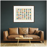 Framed abstract print featuring multicolored dots in rows on a cream background, hanging above a tan leather sofa with a small wooden coffee table and vase in a dark green room.