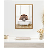 Framed canvas of a large brown bear resting its head on the edge of a white vintage clawfoot bathtub set against white paneled walls and wood flooring.