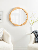 Irregular round wall mirror with natural rattan frame hung above a white table with a vase holding green branches, flanked by two cream armchairs, one with a gray throw.