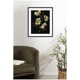 Framed floral artwork featuring cream-colored flowers with green stems on a dark background, hanging above an olive green armchair and next to a potted plant with small olives.